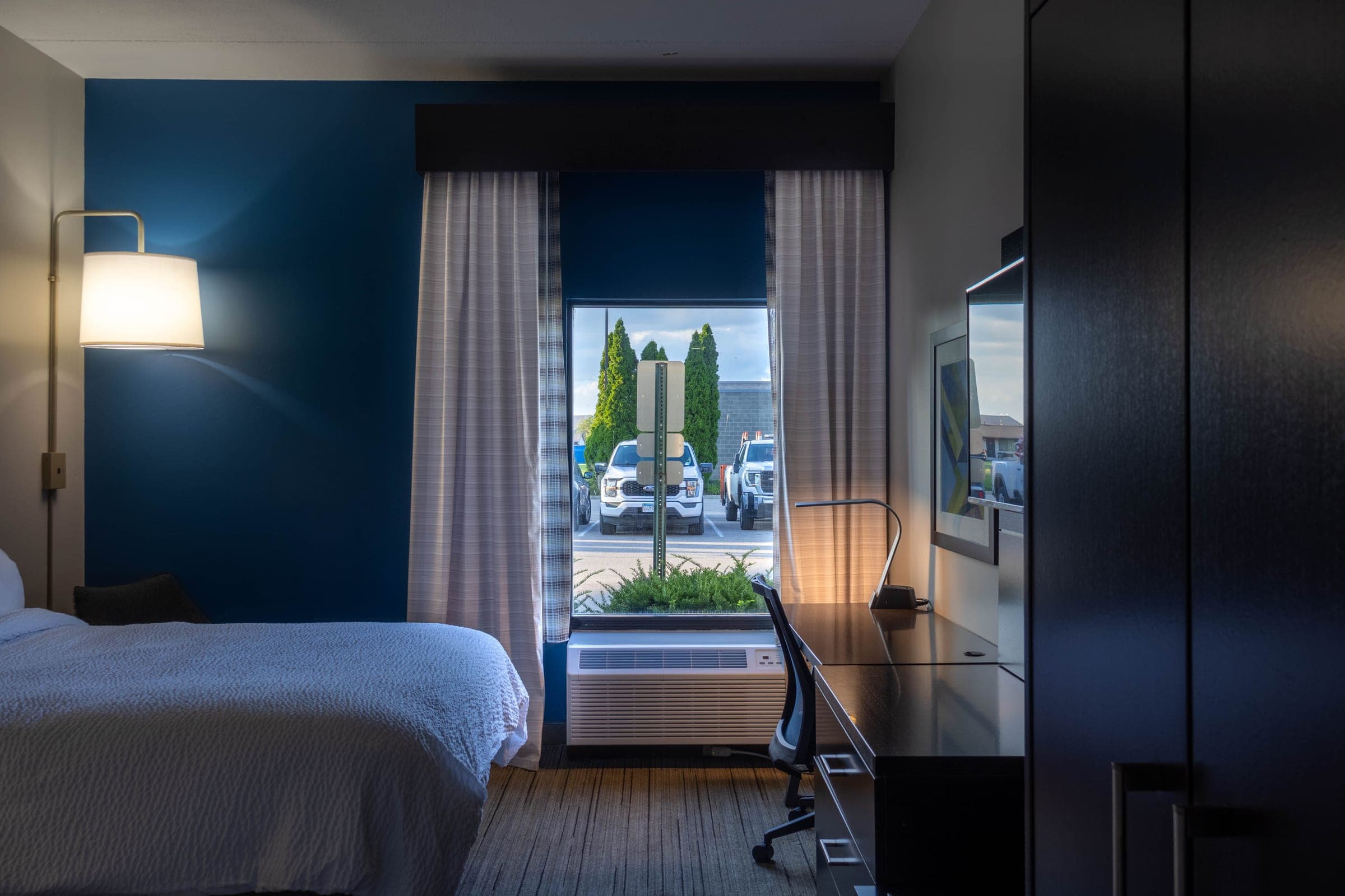 Hotel guest room with a deep blue accent wall, made bed, and a window looking out to the property entrance